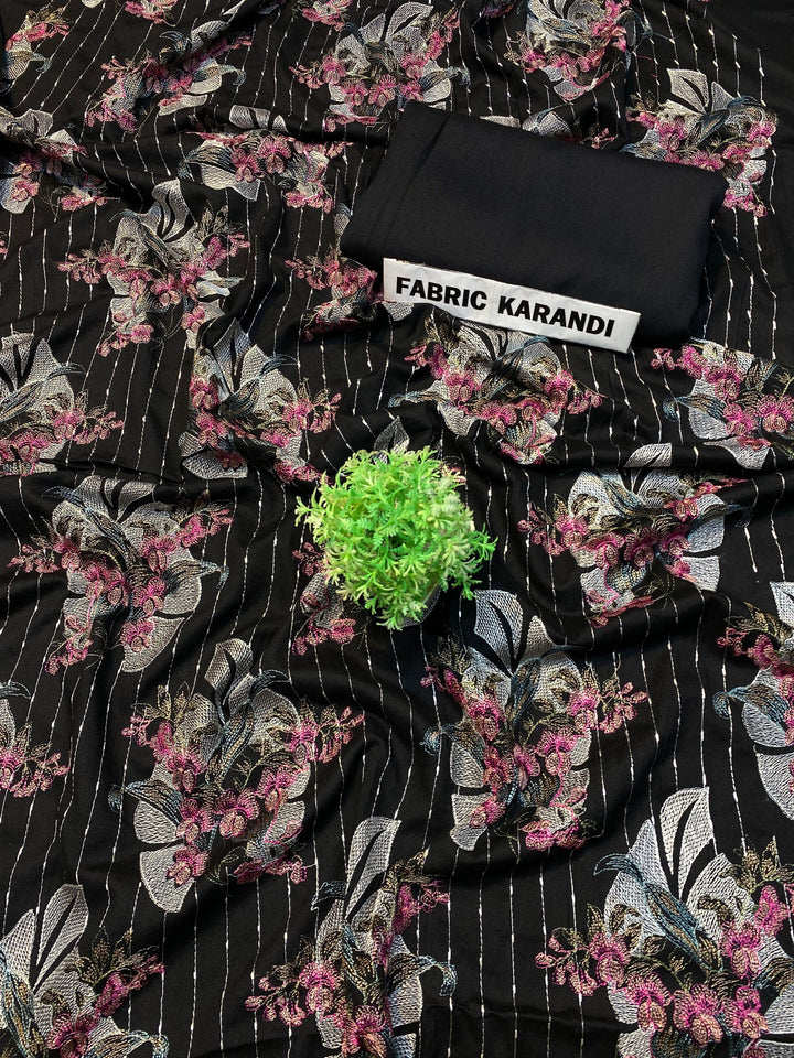 A soft pink embroidered Karandi fabric featuring floral patterns is spread out. A small green plant rests on it, and a folded piece of the same premium quality fabric lies nearby with a label reading "Golden Twilight Bareeze by ESHMEEN," perfect for the Winter Collection.