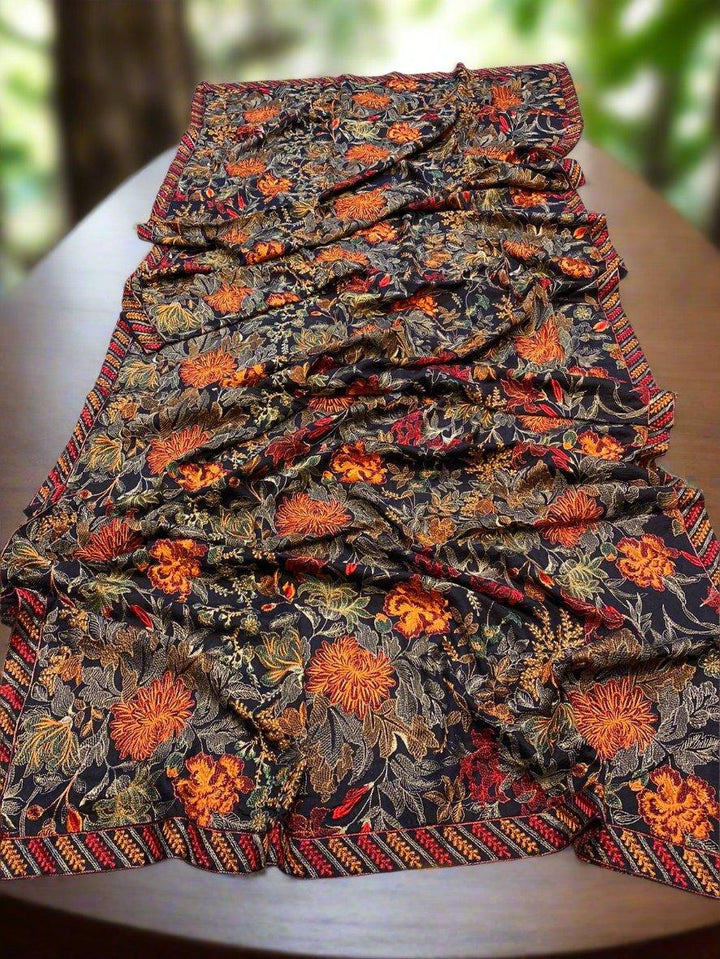 A folded Mystic Horizon Bareeze shawl by ESHMEEN lies on a wooden surface, showcasing vibrant floral patterns in shades of orange, yellow, and red against a dark background. This intricately designed heavy embroidered Karandi masterpiece features a decorative border and is available with free home delivery.