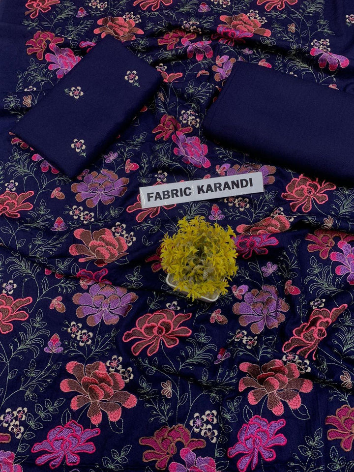 A spread labeled Bareeze Royal Flame from the ESHMEEN brand features intricate floral embroidery in pink, red, and green on a blue background. Two folded pieces of solid blue fabric rest on top, accented with a small green decorative plant—ideal for your Premium Quality Winter Collection.