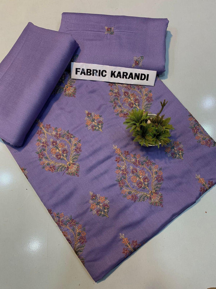 A Mystic Glory Bareeze by ESHMEEN, crafted from premium purple karandi fabric, is spread out to display its intricate multicolored patterns typical of the winter collection. Two folded pieces of matching embroidered karandi lie on top, and a small green flower bouquet graces the center. A label displays FABRIC KARANDI.