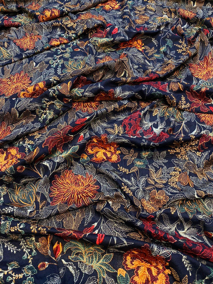 A folded Mystic Horizon Bareeze shawl by ESHMEEN lies on a wooden surface, showcasing vibrant floral patterns in shades of orange, yellow, and red against a dark background. This intricately designed heavy embroidered Karandi masterpiece features a decorative border and is available with free home delivery.