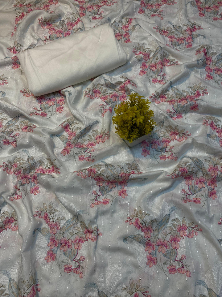 A soft pink embroidered Karandi fabric featuring floral patterns is spread out. A small green plant rests on it, and a folded piece of the same premium quality fabric lies nearby with a label reading "Golden Twilight Bareeze by ESHMEEN," perfect for the Winter Collection.