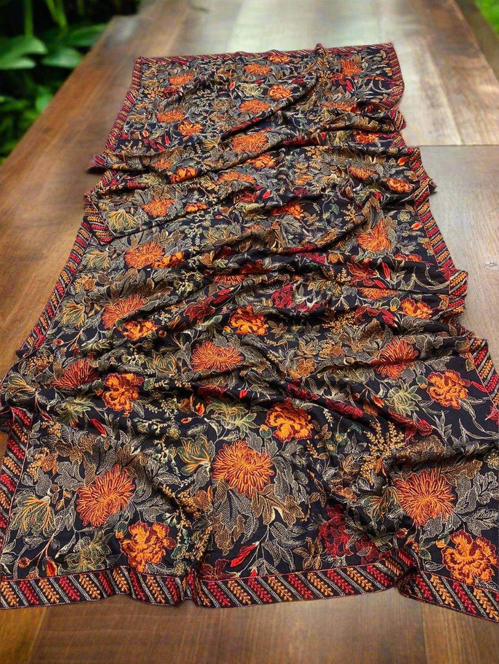 A beautifully adorned Mystic Opal Bareeze by ESHMEEN, featuring an intricate floral pattern in shades of orange, red, and gold on a dark background, is elegantly draped on a wooden table. The edges boast a geometric design that complements the floral motifs—ideal for upgrading your winter wardrobe.