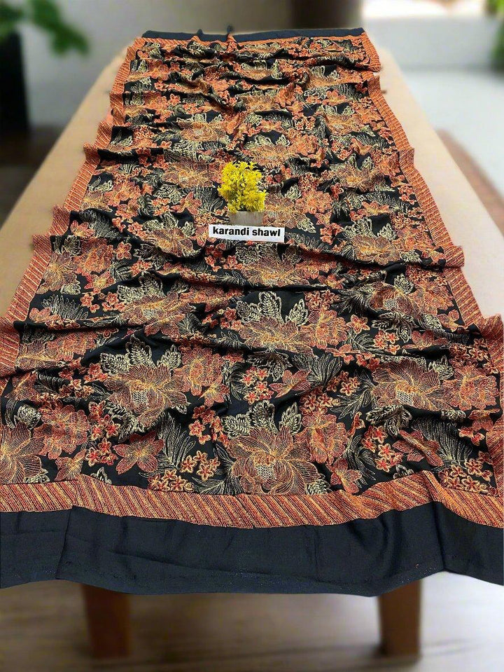 The Bareeze Radiant Charm shawl from ESHMEEN, part of the winter collection, showcases intricate floral patterns in orange and yellow tones. It is gracefully spread on a wooden table with a small yellow flower delicately placed at its center.