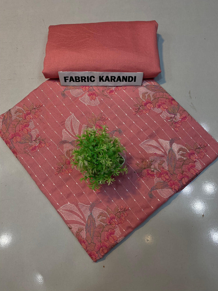 A soft pink embroidered Karandi fabric featuring floral patterns is spread out. A small green plant rests on it, and a folded piece of the same premium quality fabric lies nearby with a label reading "Golden Twilight Bareeze by ESHMEEN," perfect for the Winter Collection.