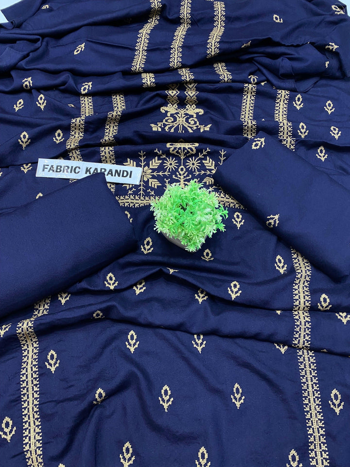 A blue fabric with intricate gold embroidery is spread out, showcasing its detailed design. A small green plant is placed on top, with a label reading Eternal Charm Bareeze from ESHMEEN's Winter Collection.