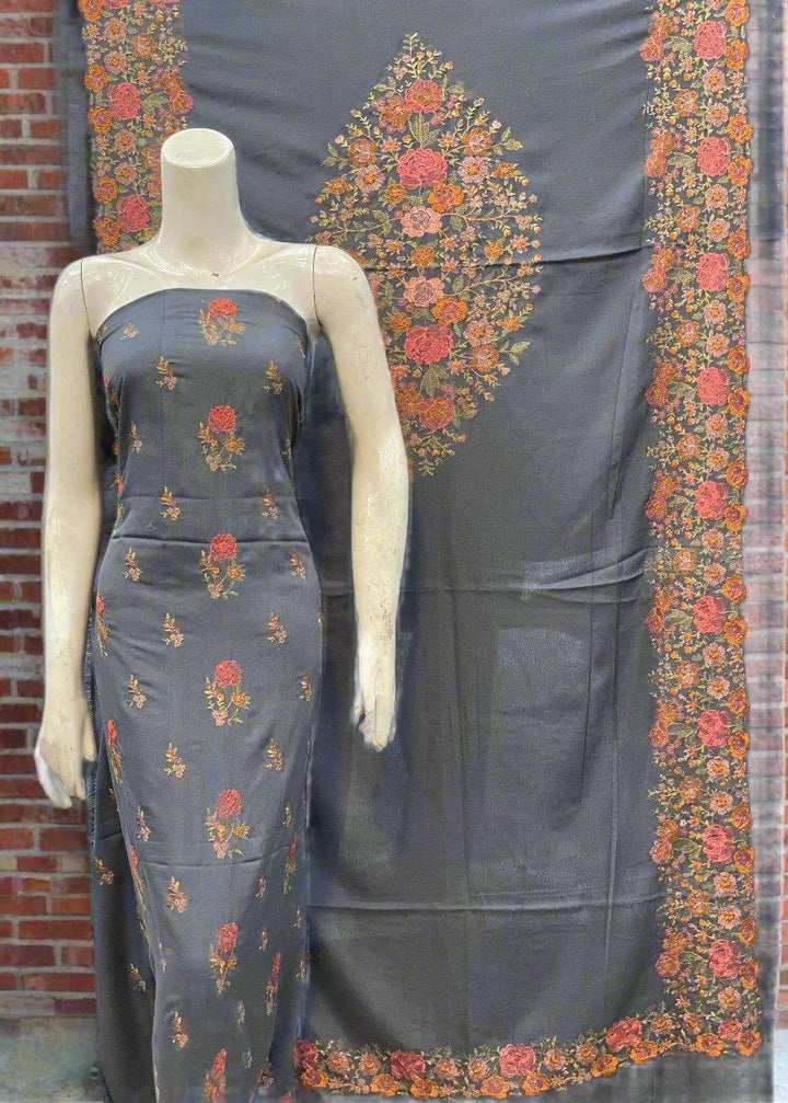 The mannequin is elegantly adorned with ESHMEEN's Velvet Dream Bareeze, featuring a gray fabric with intricate orange and red floral patterns and an embroidered Karandi texture. A matching fabric panel showcasing central and border floral motifs is displayed behind it against a brick wall, capturing the essence of this exquisite winter collection.