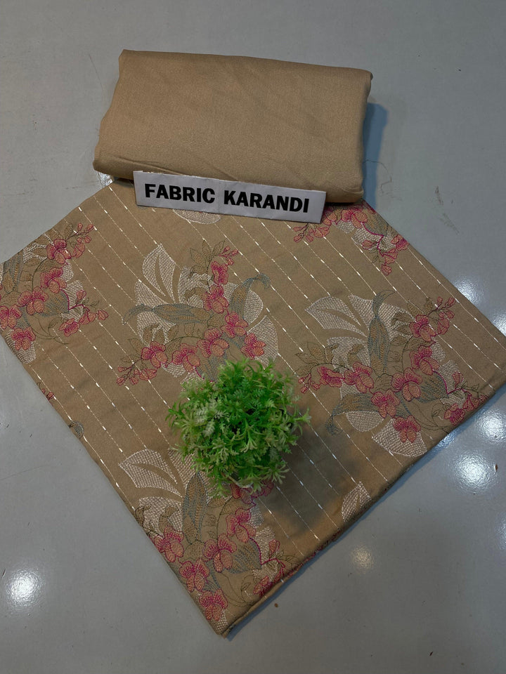 A soft pink embroidered Karandi fabric featuring floral patterns is spread out. A small green plant rests on it, and a folded piece of the same premium quality fabric lies nearby with a label reading "Golden Twilight Bareeze by ESHMEEN," perfect for the Winter Collection.