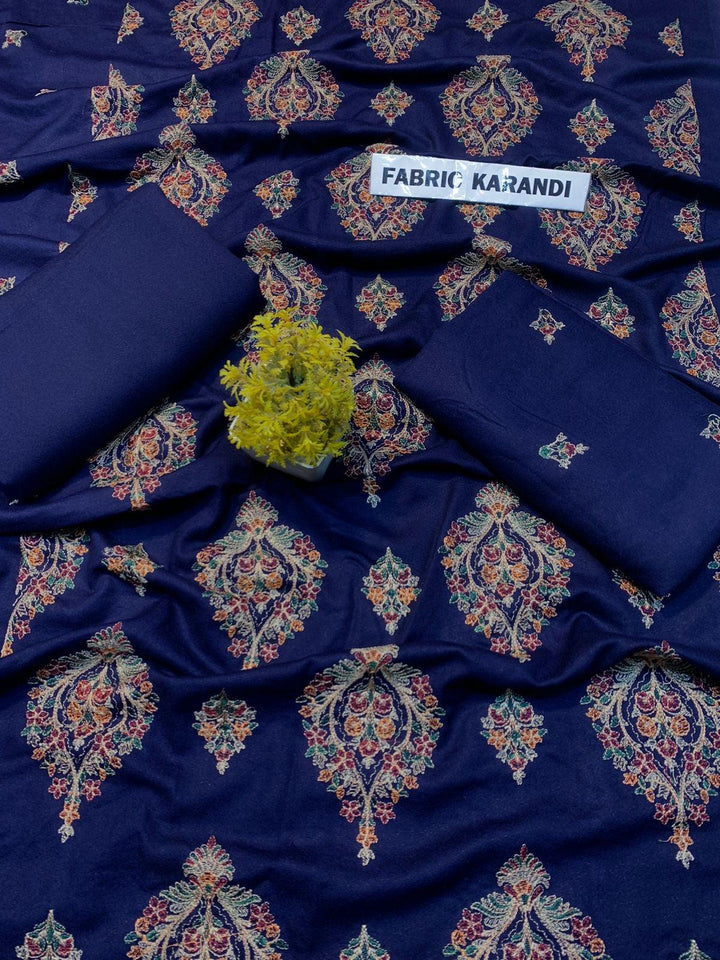 A Mystic Glory Bareeze by ESHMEEN, crafted from premium purple karandi fabric, is spread out to display its intricate multicolored patterns typical of the winter collection. Two folded pieces of matching embroidered karandi lie on top, and a small green flower bouquet graces the center. A label displays FABRIC KARANDI.