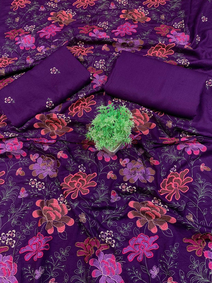 A spread labeled Bareeze Royal Flame from the ESHMEEN brand features intricate floral embroidery in pink, red, and green on a blue background. Two folded pieces of solid blue fabric rest on top, accented with a small green decorative plant—ideal for your Premium Quality Winter Collection.