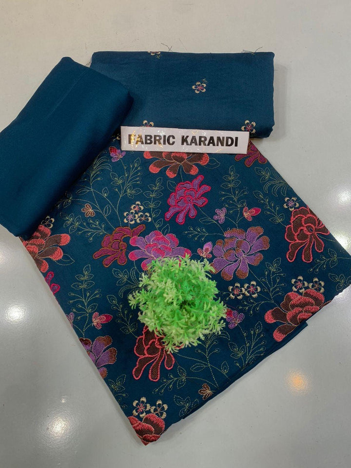 A spread labeled Bareeze Royal Flame from the ESHMEEN brand features intricate floral embroidery in pink, red, and green on a blue background. Two folded pieces of solid blue fabric rest on top, accented with a small green decorative plant—ideal for your Premium Quality Winter Collection.