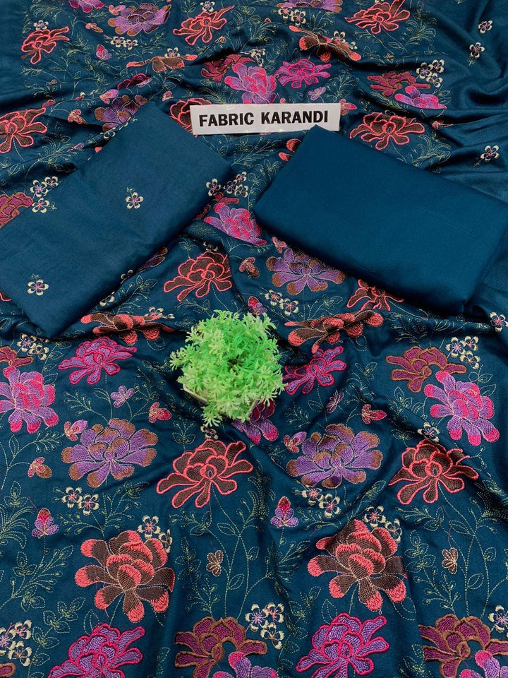 A spread labeled Bareeze Royal Flame from the ESHMEEN brand features intricate floral embroidery in pink, red, and green on a blue background. Two folded pieces of solid blue fabric rest on top, accented with a small green decorative plant—ideal for your Premium Quality Winter Collection.