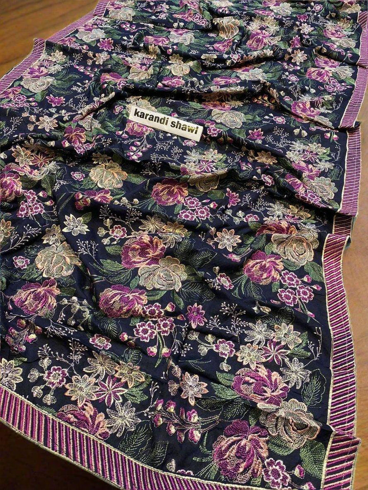 An Imperial Charm Bareeze by ESHMEEN, an ideal addition to your winter wardrobe, is elegantly laid over a wooden table. The floral design in pink, green, and beige stands out against the dark background with a striped edge. Softly blurred lush greenery provides the perfect backdrop. Relish free home delivery with this exquisite piece.