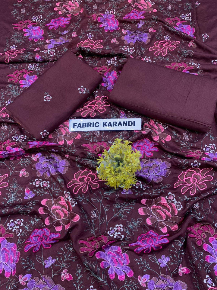 A spread labeled Bareeze Royal Flame from the ESHMEEN brand features intricate floral embroidery in pink, red, and green on a blue background. Two folded pieces of solid blue fabric rest on top, accented with a small green decorative plant—ideal for your Premium Quality Winter Collection.