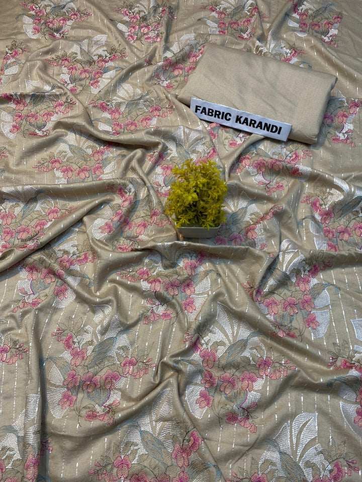 A soft pink embroidered Karandi fabric featuring floral patterns is spread out. A small green plant rests on it, and a folded piece of the same premium quality fabric lies nearby with a label reading "Golden Twilight Bareeze by ESHMEEN," perfect for the Winter Collection.