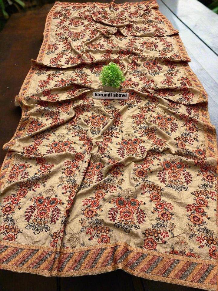 A beige shawl adorned with intricate red and brown floral patterns is elegantly displayed on a wooden surface, featuring a striped border and accentuated by a small green plant placed atop. Experience the essence of the ESHMEEN Noble Essence Bareeze from the winter collection. Enjoy free home delivery today!