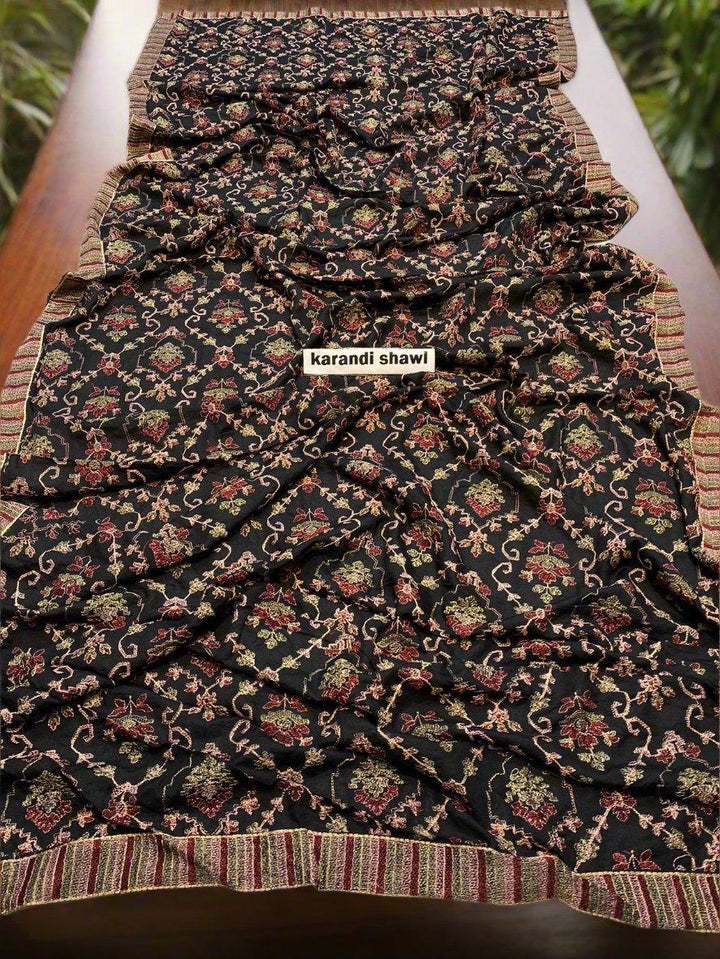 A black Regal Dawn Bareeze Shawl from ESHMEEN, featuring intricate floral patterns in red, green, and gold, is elegantly displayed on a flat wooden surface. This shawl, part of the winter collection, showcases a striped border in coordinating colors. A small label indicates "karandi shawl." Enjoy free home delivery!