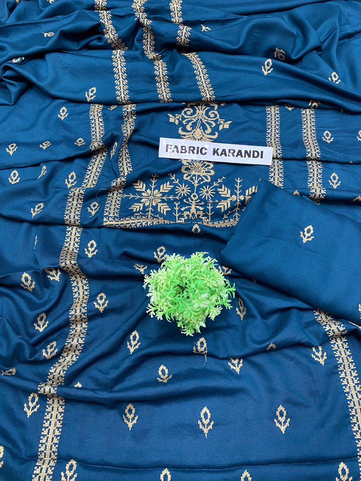 A blue fabric with intricate gold embroidery is spread out, showcasing its detailed design. A small green plant is placed on top, with a label reading Eternal Charm Bareeze from ESHMEEN's Winter Collection.