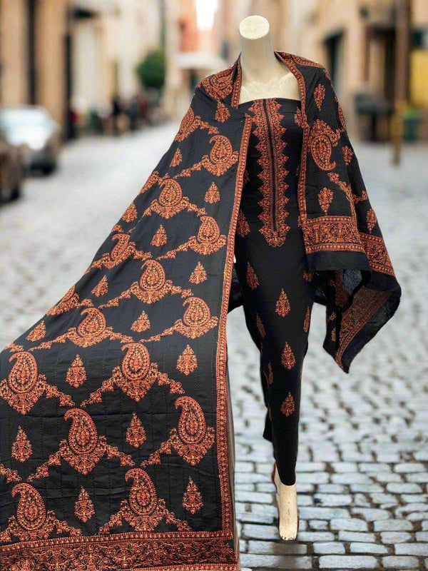 On a cobblestone street, a mannequin showcases the ESHMEEN Bareeze Mystic Pearl outfit—a viscose-embroidered karandi traditional ensemble adorned with intricate patterns. The winter collection includes a black and red shawl beautifully draped over its shoulders, set against a blurred street scene backdrop.