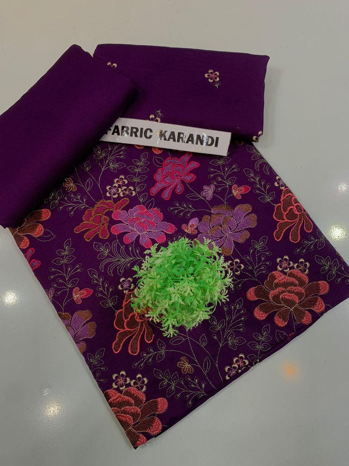 A spread labeled Bareeze Royal Flame from the ESHMEEN brand features intricate floral embroidery in pink, red, and green on a blue background. Two folded pieces of solid blue fabric rest on top, accented with a small green decorative plant—ideal for your Premium Quality Winter Collection.