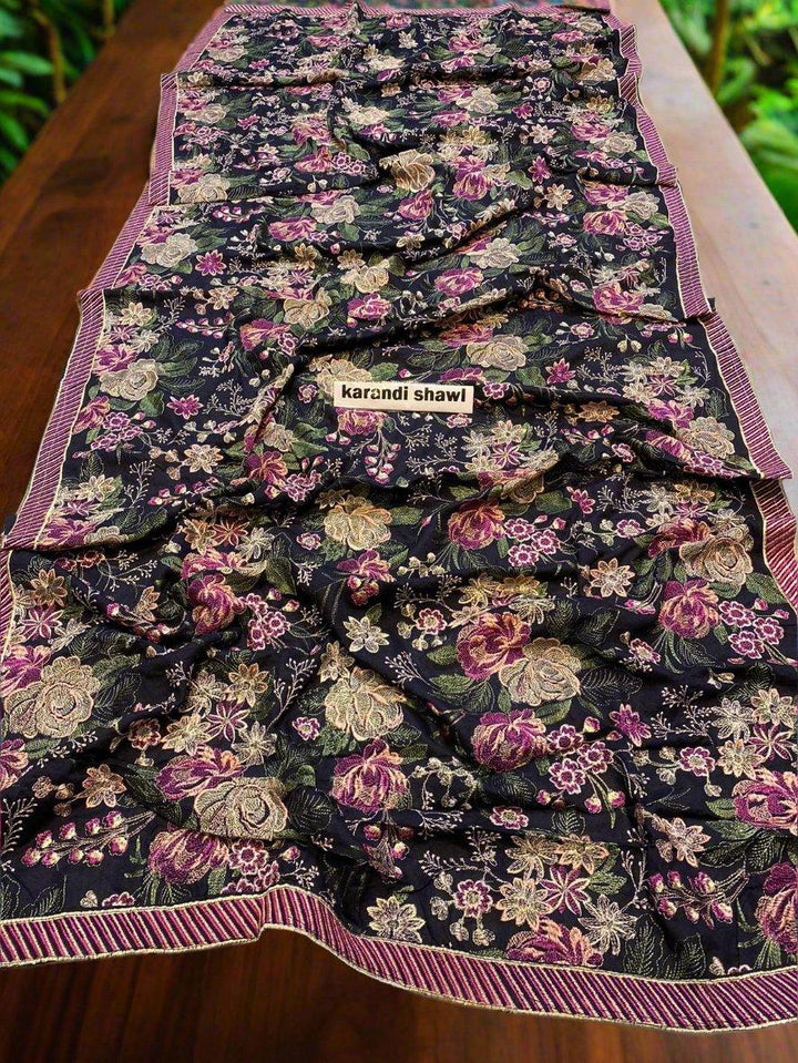 An Imperial Charm Bareeze by ESHMEEN, an ideal addition to your winter wardrobe, is elegantly laid over a wooden table. The floral design in pink, green, and beige stands out against the dark background with a striped edge. Softly blurred lush greenery provides the perfect backdrop. Relish free home delivery with this exquisite piece.