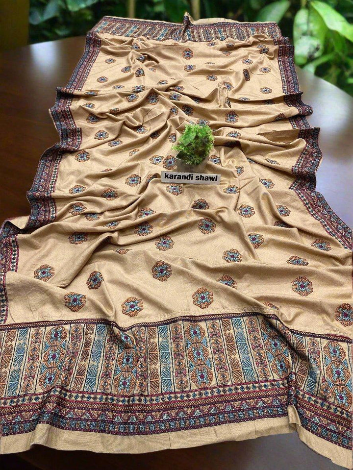 On a wooden table lies a textured beige shawl featuring intricate blue and burgundy floral patterns from ESHMEEN's Crimson Veil Bareeze collection. A small green plant, labeled as part of your perfect winter wardrobe, is placed on the fabric. Enjoy free home delivery with this exquisite piece.