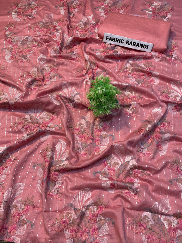 A soft pink embroidered Karandi fabric featuring floral patterns is spread out. A small green plant rests on it, and a folded piece of the same premium quality fabric lies nearby with a label reading "Golden Twilight Bareeze by ESHMEEN," perfect for the Winter Collection.