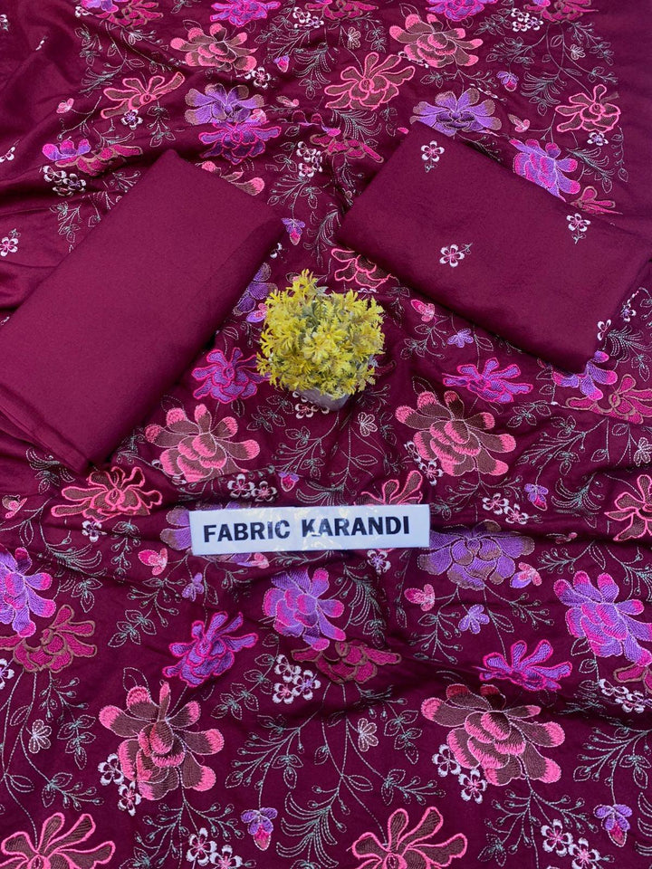 A spread labeled Bareeze Royal Flame from the ESHMEEN brand features intricate floral embroidery in pink, red, and green on a blue background. Two folded pieces of solid blue fabric rest on top, accented with a small green decorative plant—ideal for your Premium Quality Winter Collection.