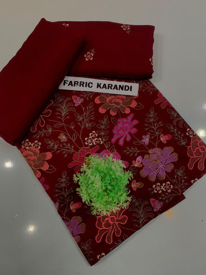 A spread labeled Bareeze Royal Flame from the ESHMEEN brand features intricate floral embroidery in pink, red, and green on a blue background. Two folded pieces of solid blue fabric rest on top, accented with a small green decorative plant—ideal for your Premium Quality Winter Collection.