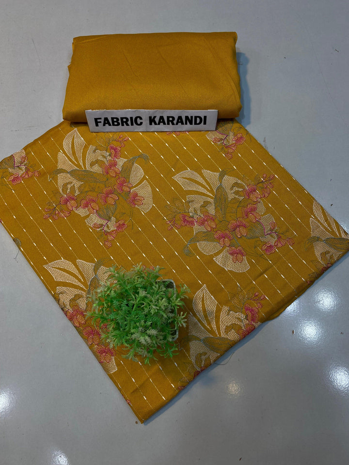 A soft pink embroidered Karandi fabric featuring floral patterns is spread out. A small green plant rests on it, and a folded piece of the same premium quality fabric lies nearby with a label reading "Golden Twilight Bareeze by ESHMEEN," perfect for the Winter Collection.